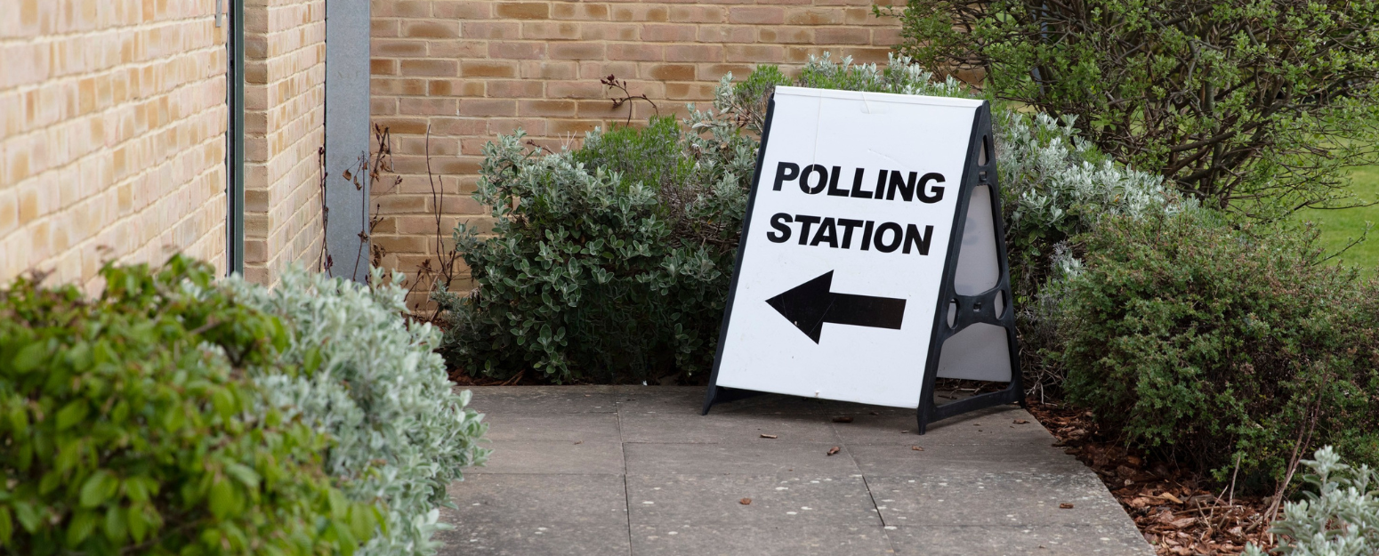 A‑frame sign on a pathway next to a brick building, reading ‘Polling Station’ with a left‑pointing arrow, surrounded by shrubs.