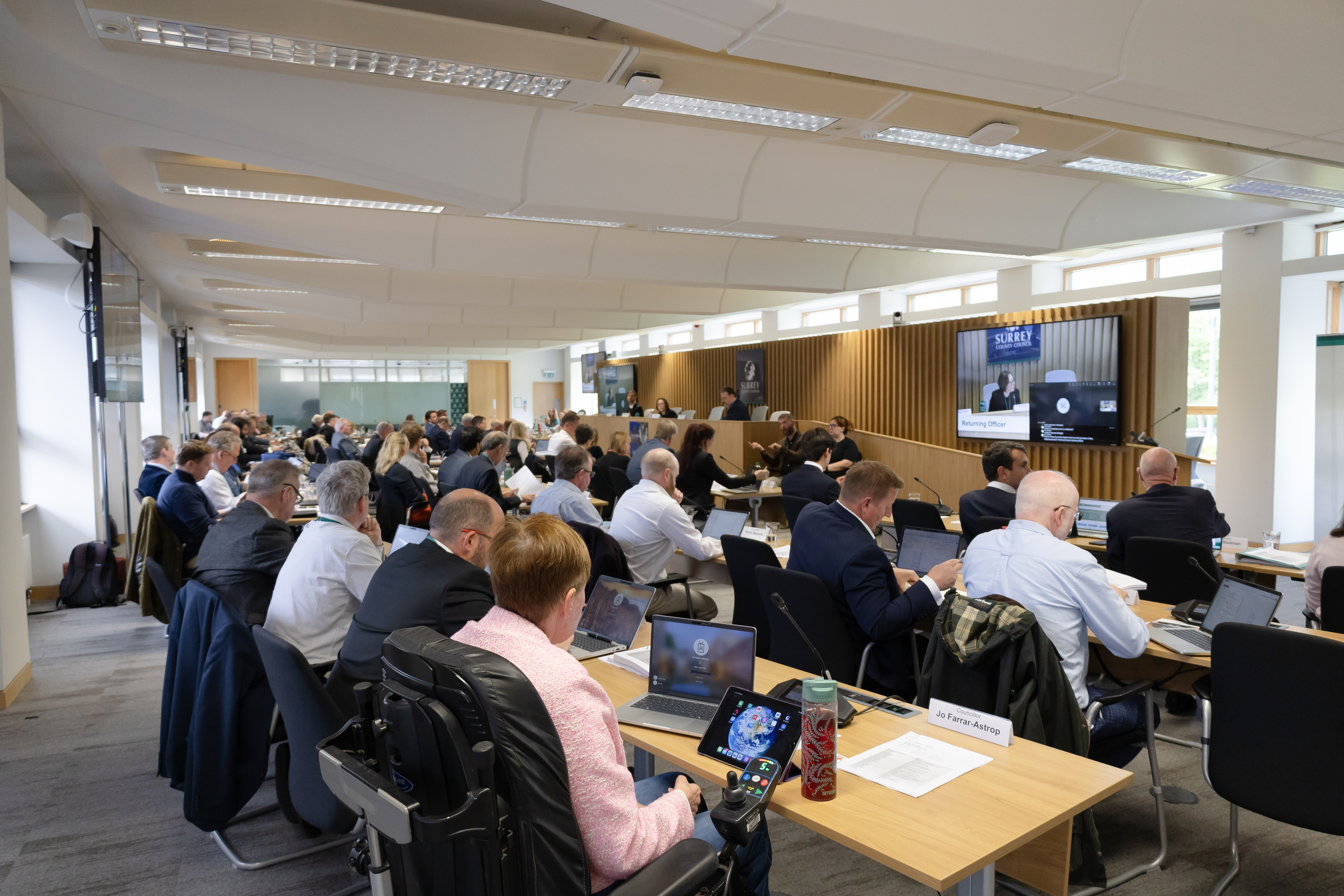 A formal meeting in progress with attendees seated around tables, some participating via video conference on a large screen.